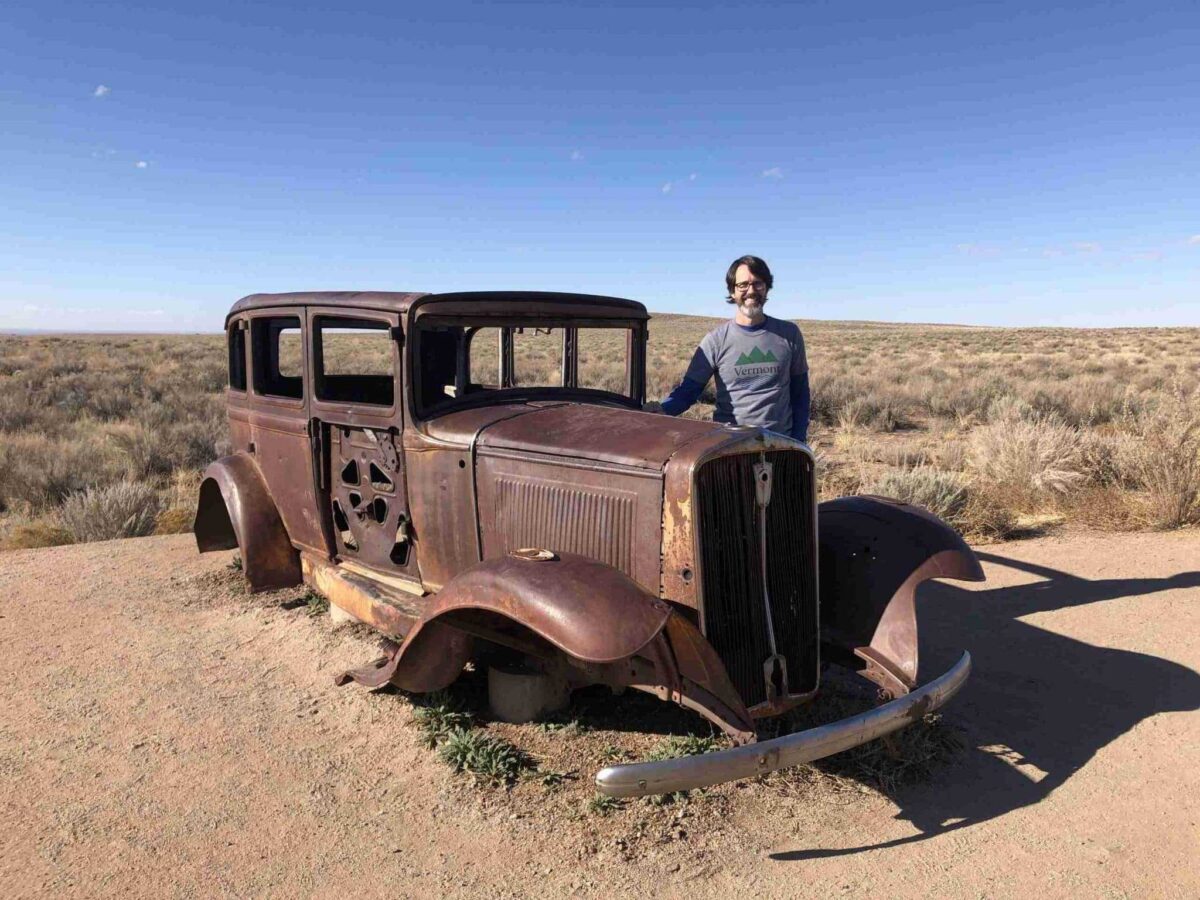 A man stands next to a rusted car.