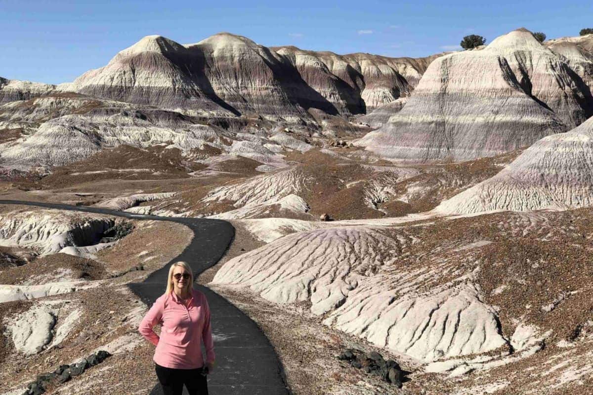 A woman stands on a paved path by colorful, striped mounds - Blue Mesa Trail in Petrified Forest National Park.