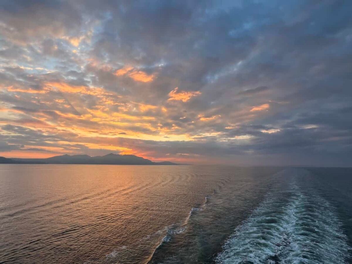 The sun sets behind mountains on the horizon as waves form behind a cruise ship.