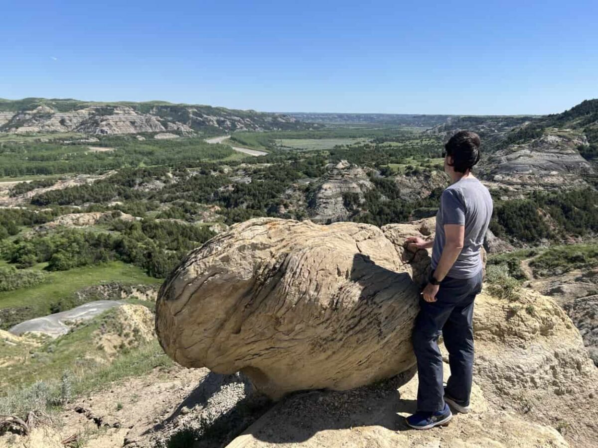 A man stands behind a big rock and enjoys the view of badlands below.