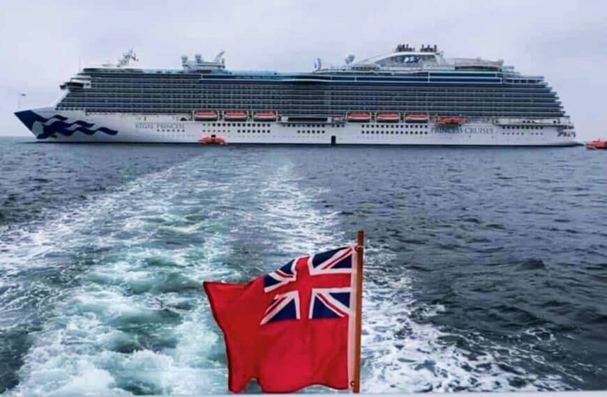 A British flag waves in the wind from the back of a tender that pulls away from a cruise ship.