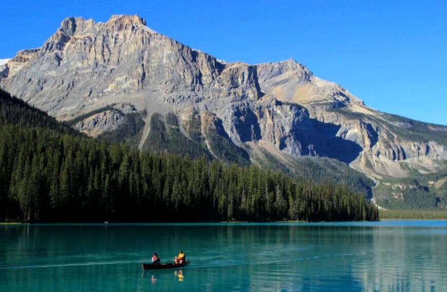Two people row a canoe in a beautiful blue lake with a towering mountain in the background