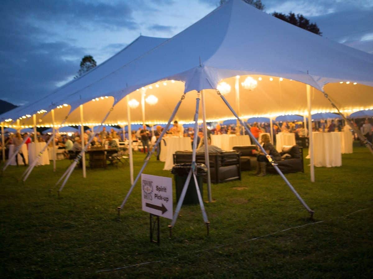 People enjoy food and beverages under a large white tent with string lights.
