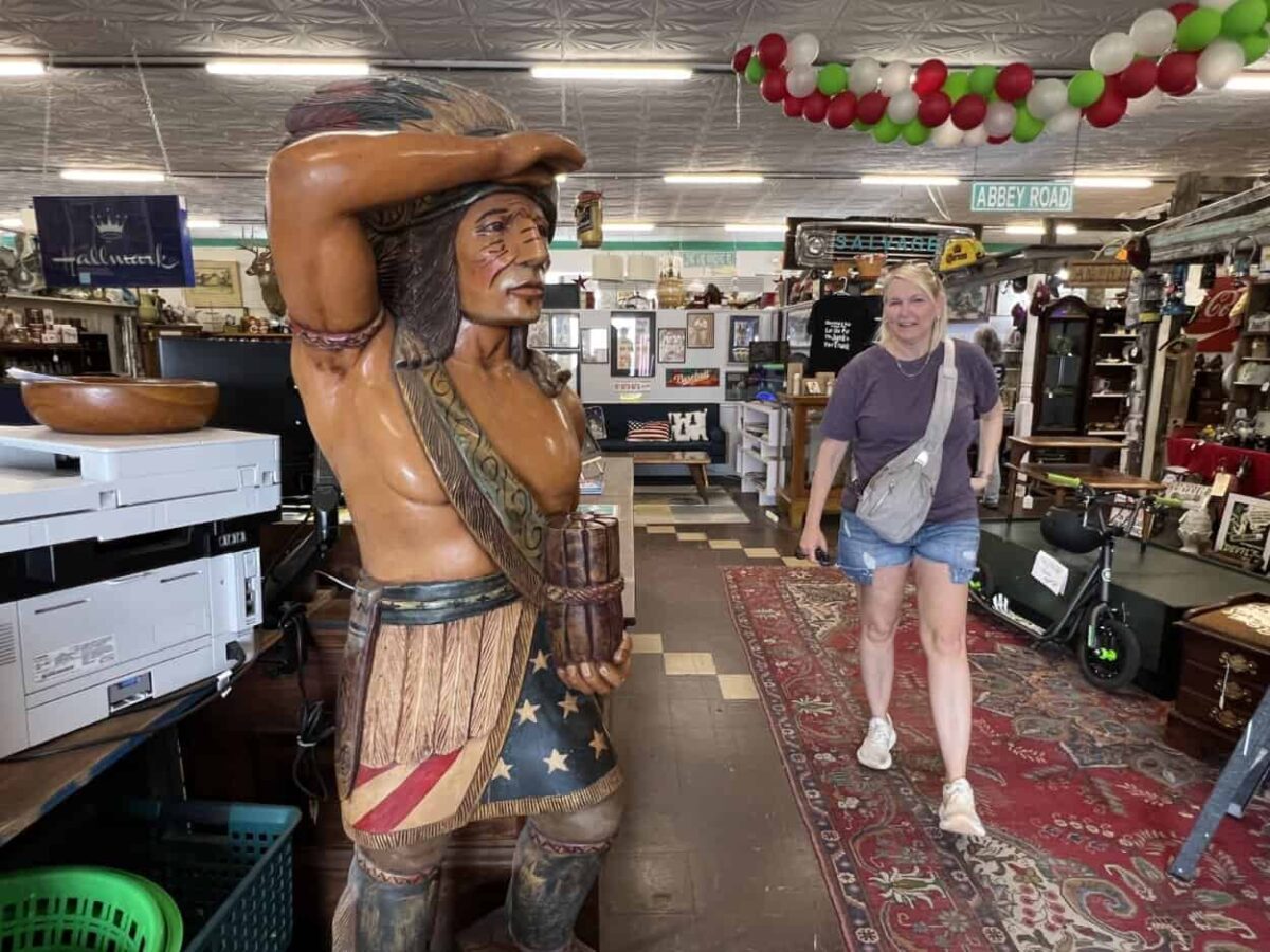 A life-sized wooden carved Indian stands in the front of an antique mall as a woman walks toward the front of the store.