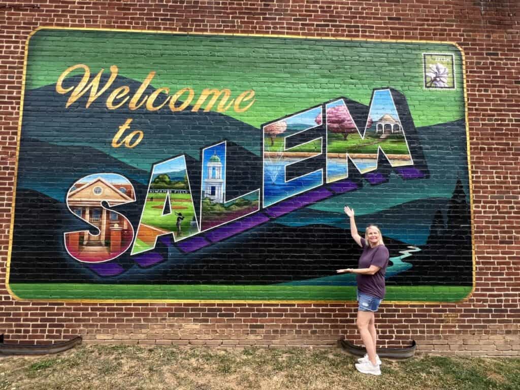 A woman stands in front of a brick wall with a colorful painted sign that ways "Welcome to Salem"