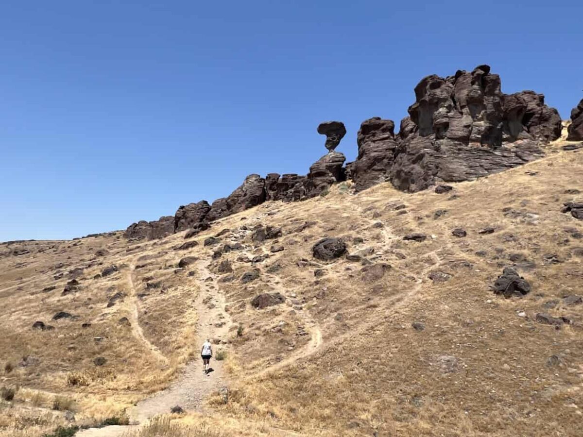 A woman hikes a trail up a hill topped with a large, brown rock formation.