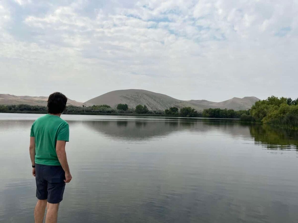 A man stands at the foot of a lake where large sand dunes rise on the horizon on the other side of the lake.