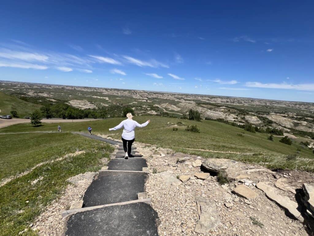 A woman walks down steps along a paved path with views of badlands in the distance