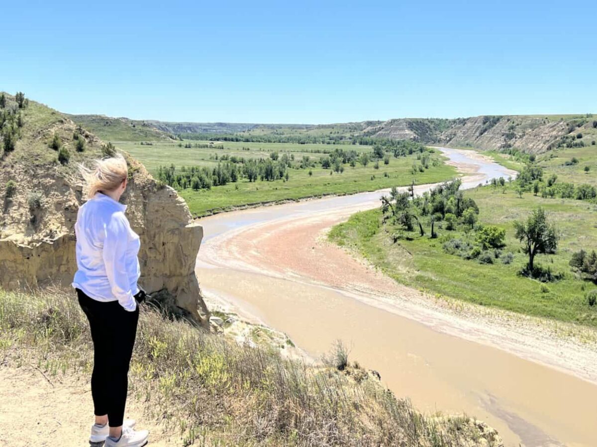 A woman stands at the edge of a cliff to look at a river winding through a valley below