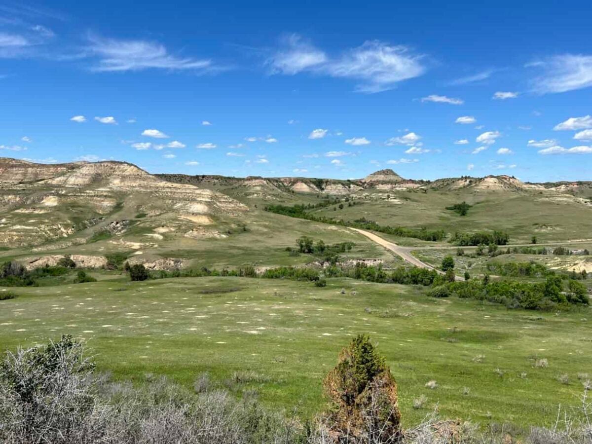 A vast field with grasslands and badlands sits under a wide open blue sky with a few white clouds