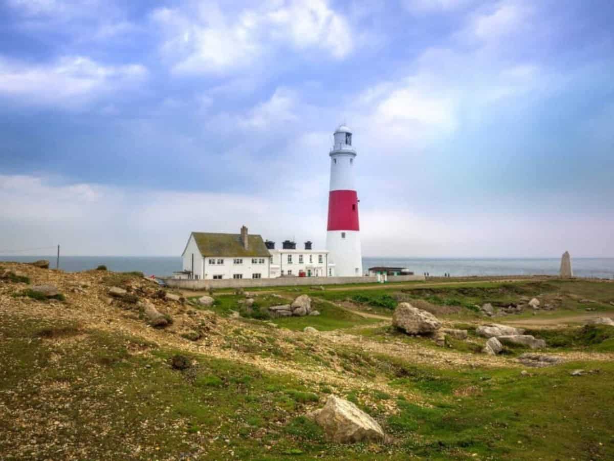 A white lighthouse with a red stripe stands across a field with green grass and brown boulders with water in the background.
