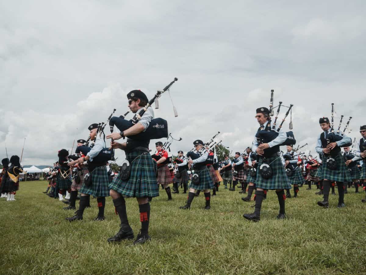 Men dressed in kilts march across a grassy field playing bagpipes