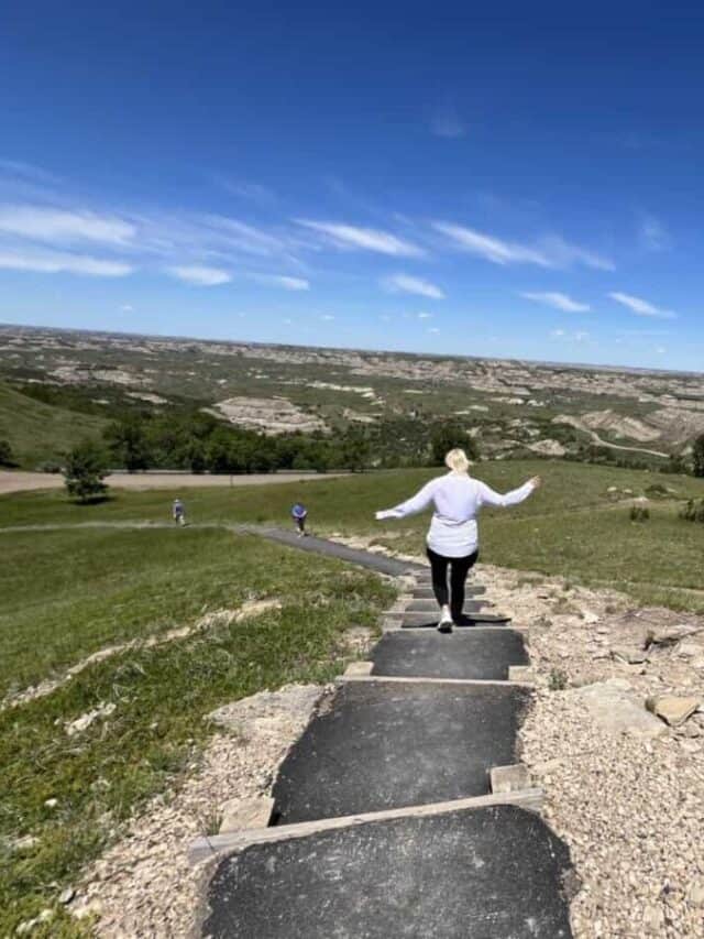 A woman walks down steps along a paved path with views of badlands in the distance