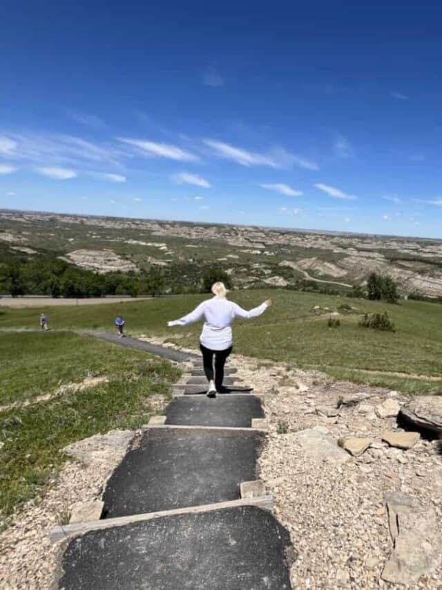 A woman walks down steps along a paved path with views of badlands in the distance