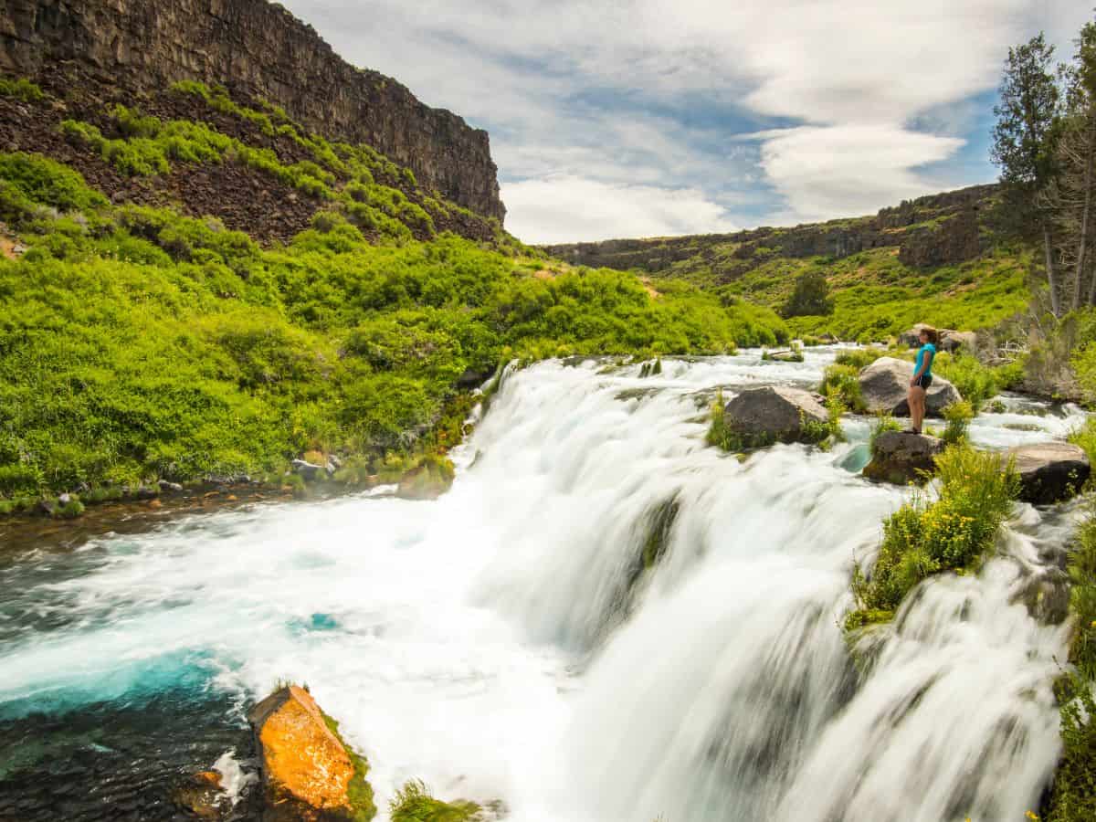 A woman stands on a rock at the top of a waterfall