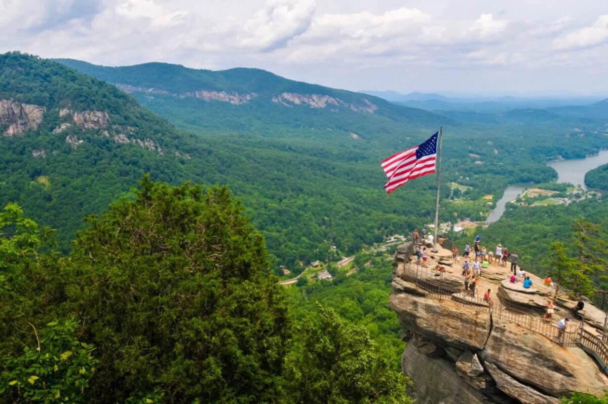 People stand on a large rock with the United States flag overlooking a deep valley with a river.