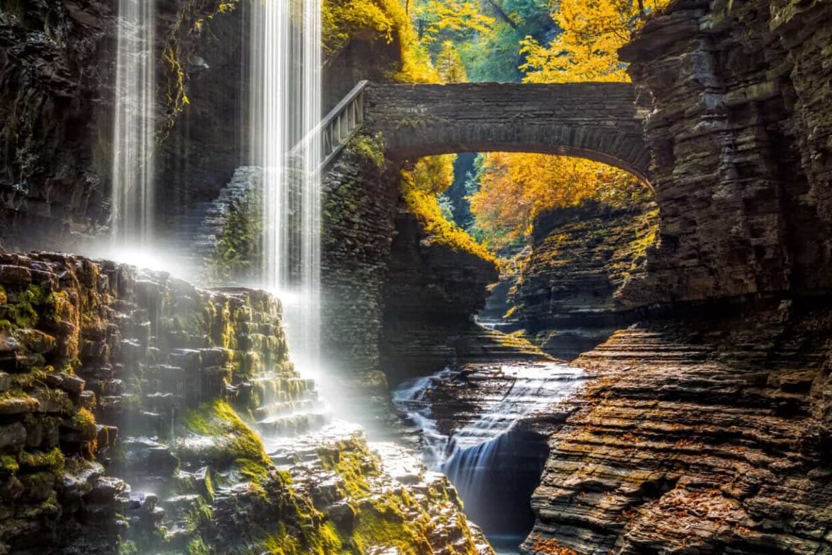 Water spills over a cliff with a stone bridge and trees with yellow leaves in the background.