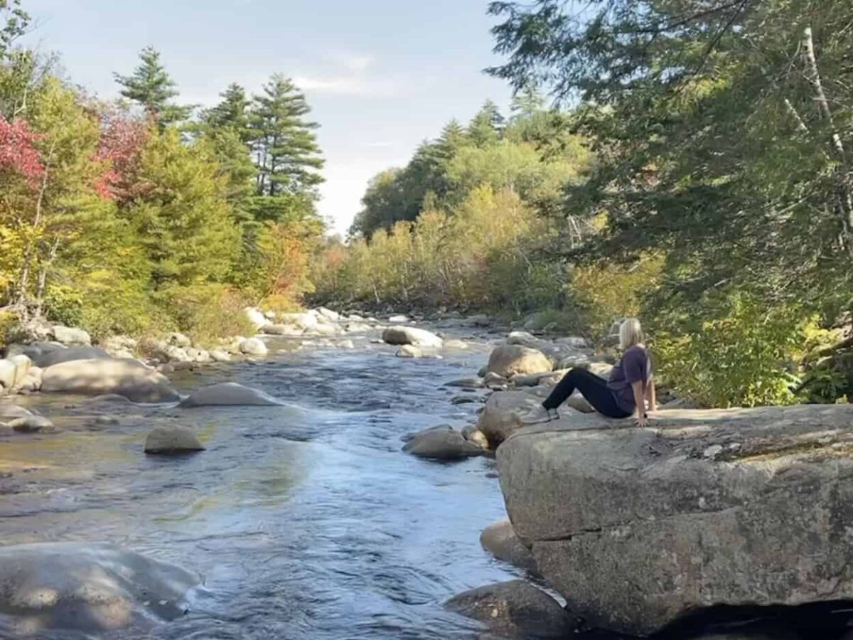 A woman sits on a large rock adjacent to a flowing river.