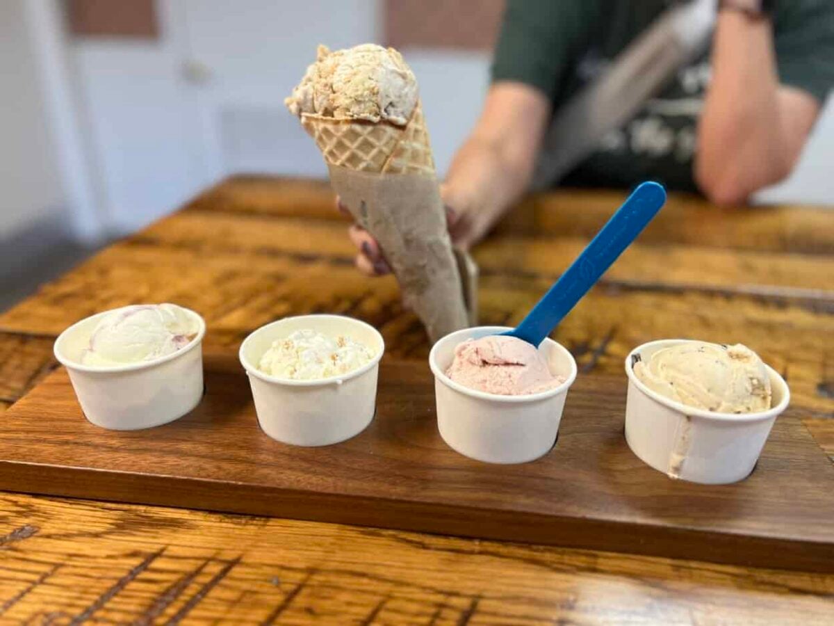 A board holds four cups of ice cream and a woman holds a waffle cone with ice cream.