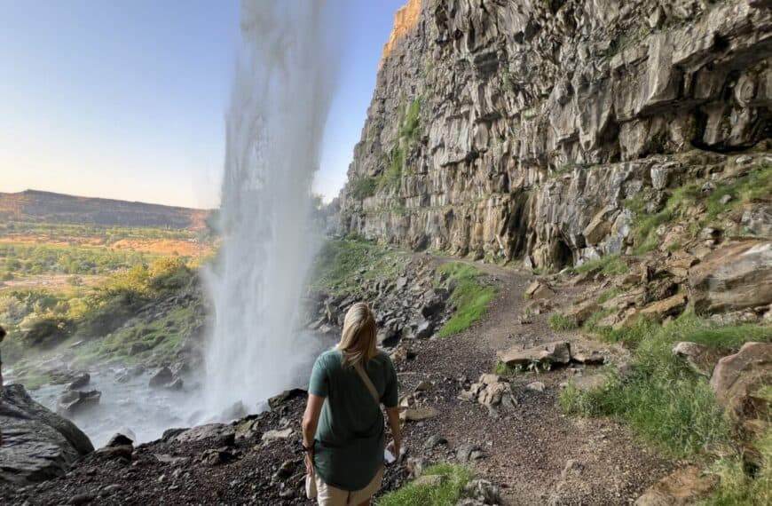A woman peers out from behind a waterfall.