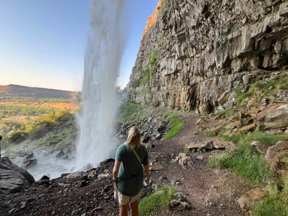 A woman peers out from behind a waterfall.
