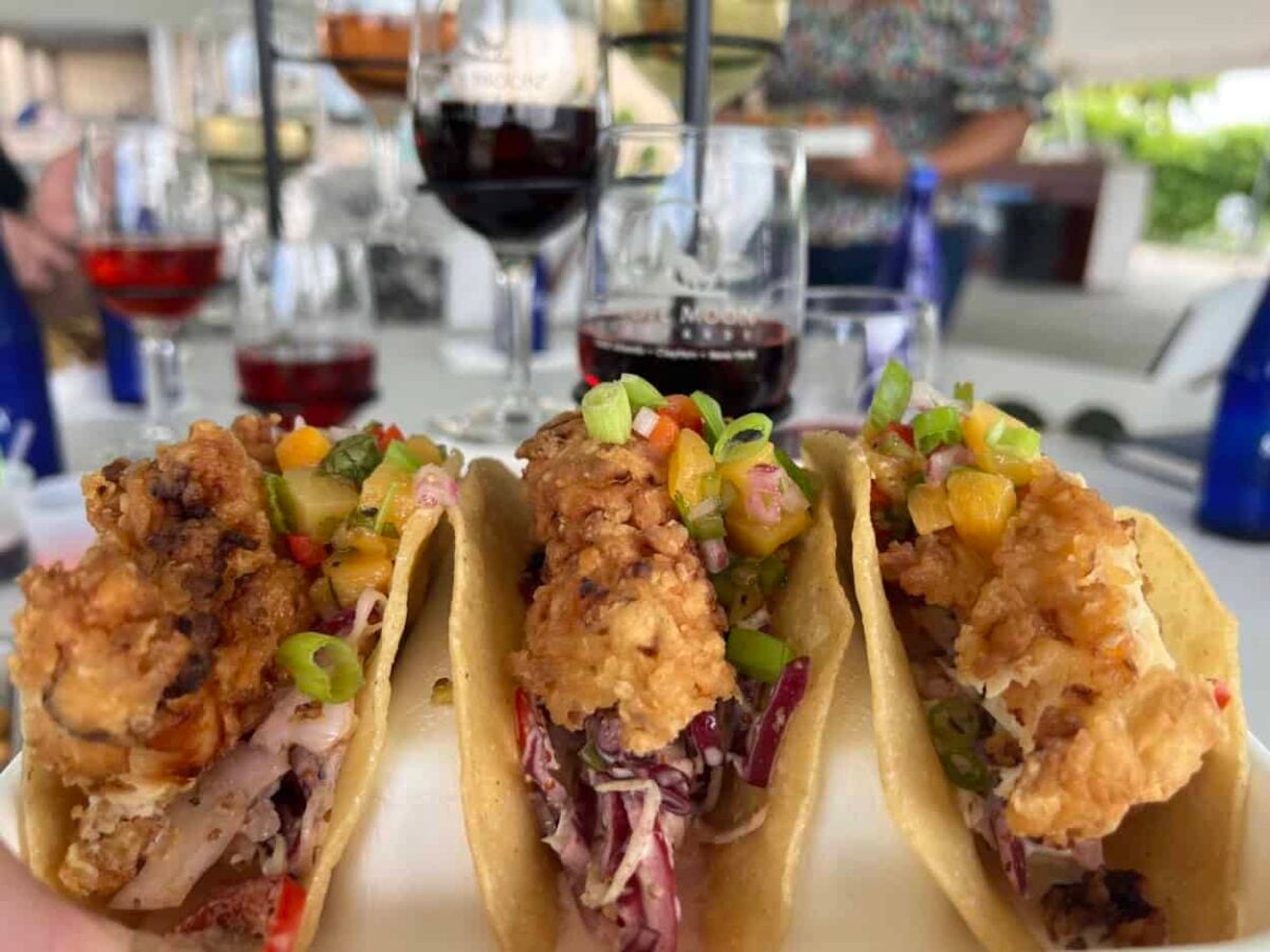 Tacos filled with slaw, fried chicken, and jalapeño peach salsa sit on a table with glasses of wine in the background.