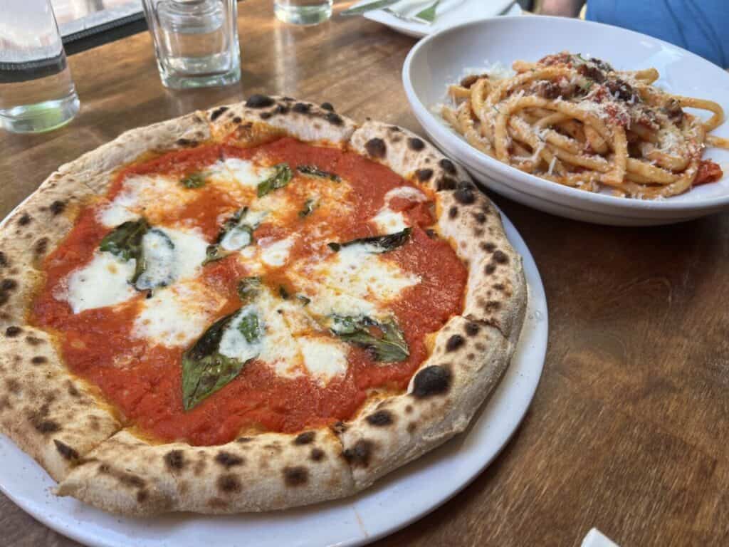 A bowl of pasta and a plate with pizza sit on a wooden table