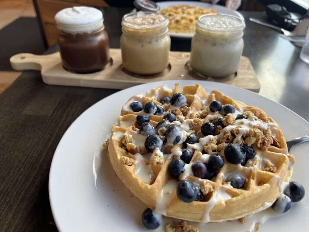 A coffee flight and a plate with a waffles topped with blueberries sit on a table.