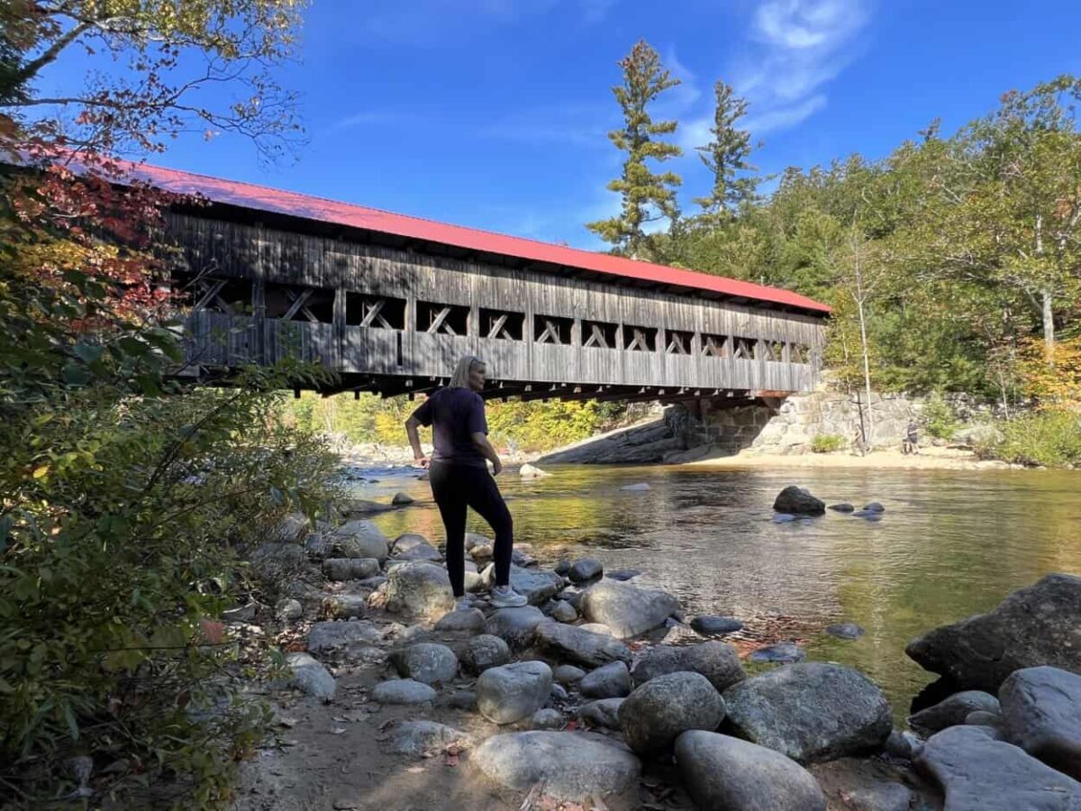 A woman steps across rocks to get a closer look at a wooden covered bridge across a river.