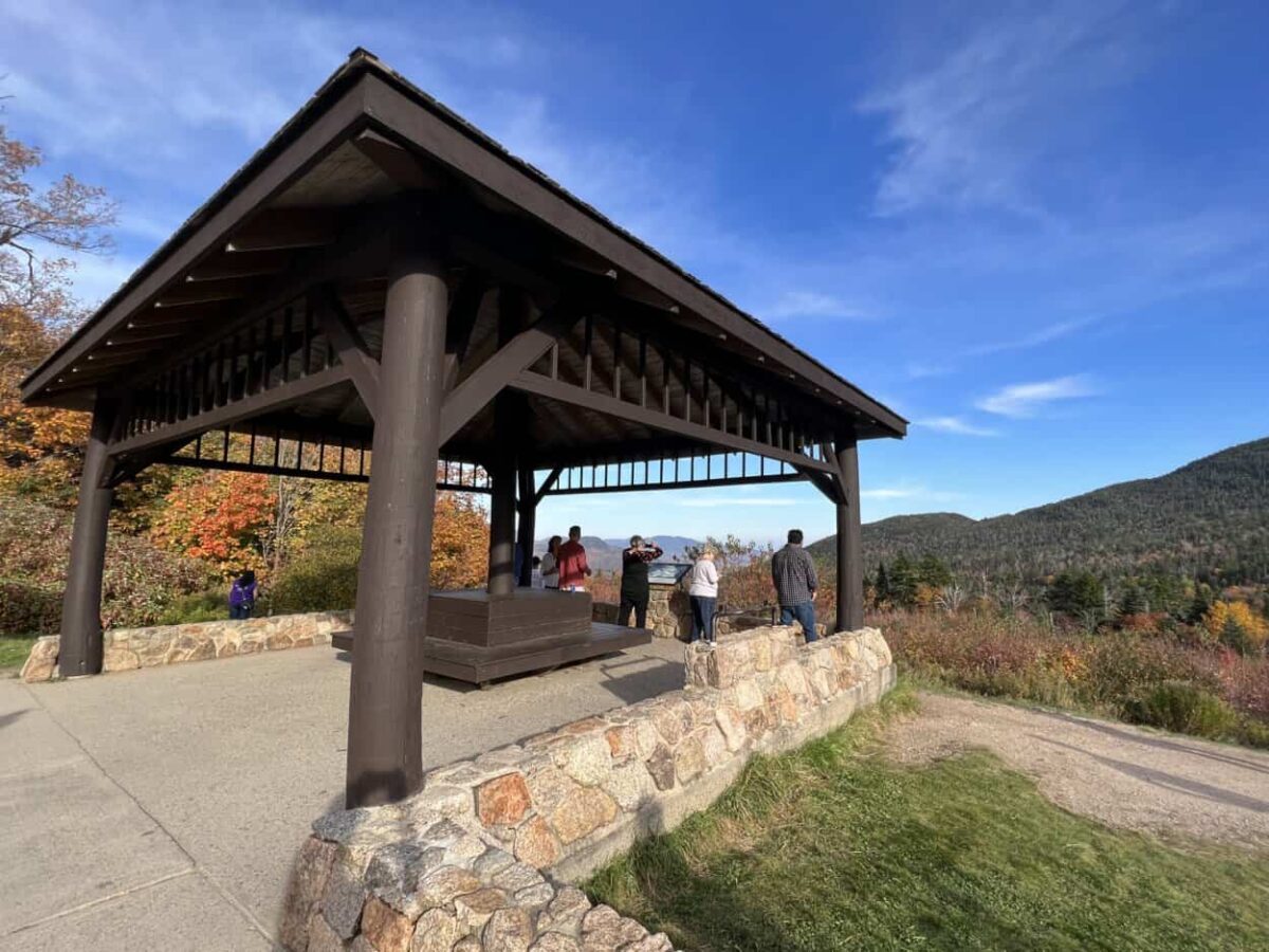 People stand under a shelter while looking at fall foliage on a mountain in the distance.