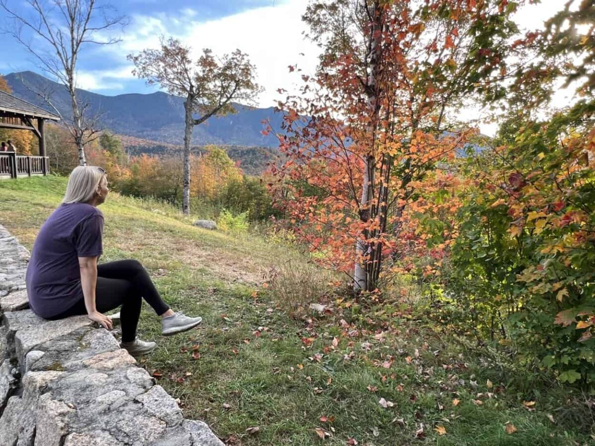 A woman sits on a stone wall while looking at mountains on the horizon.