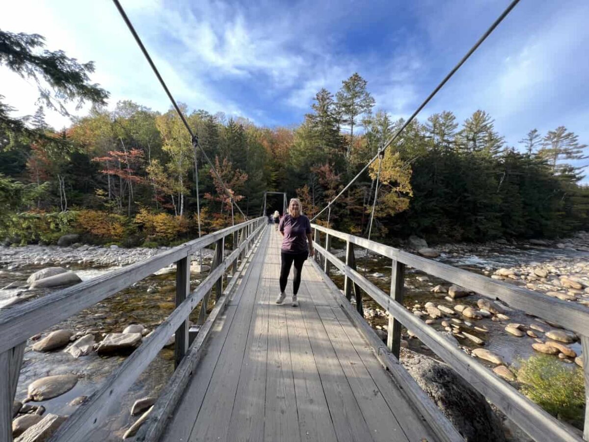 A woman stands on a wooden suspension bridge that crosses a river with many rocks.