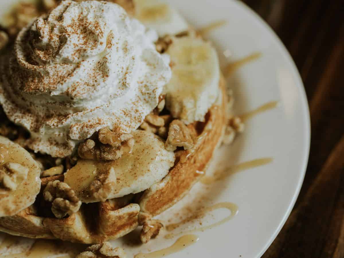 A waffle covered with banana slices, nuts, whipped cream, and cinnamon sits on a white plate