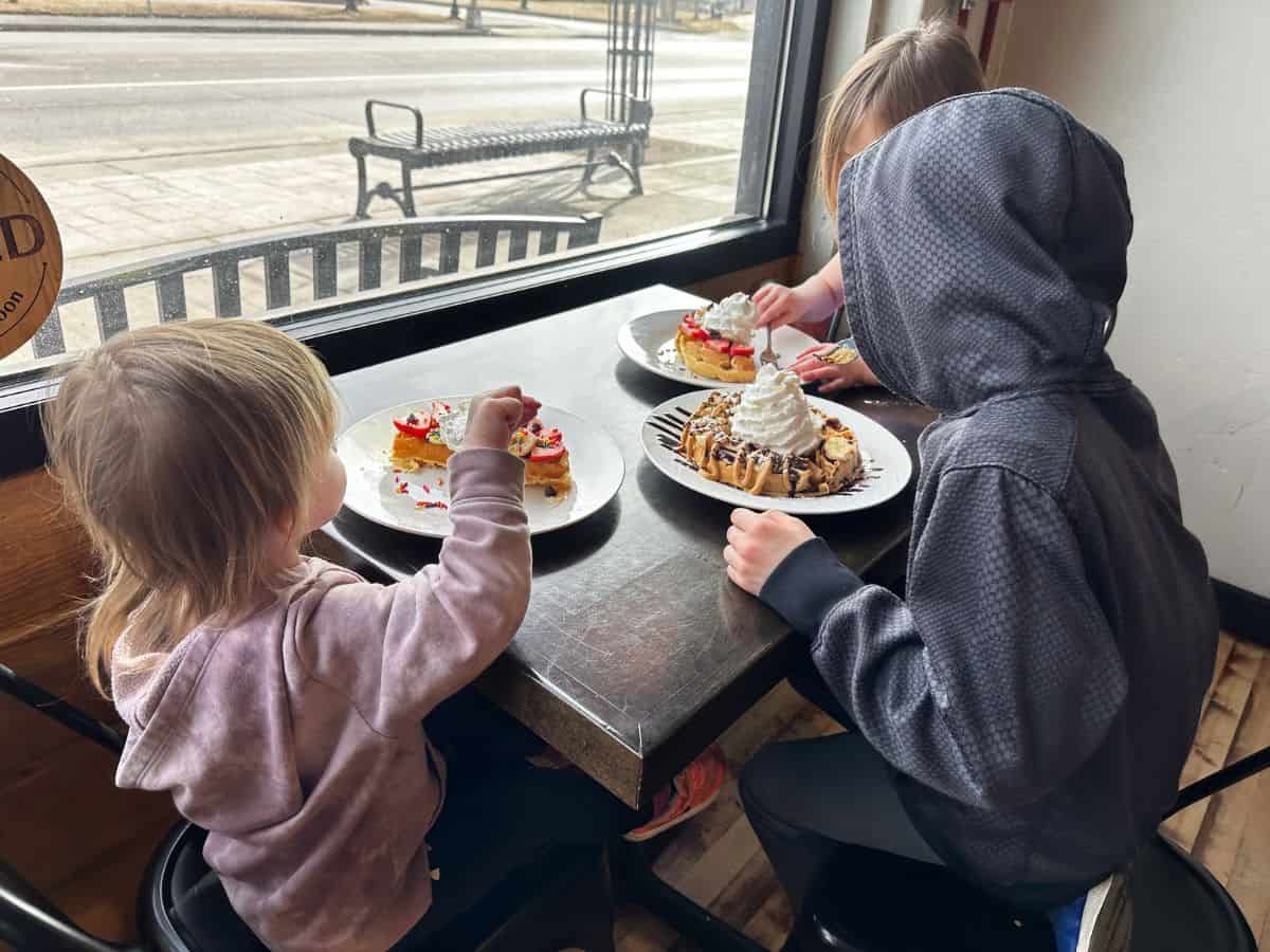 Three children sit at a table eating waffles topped with whipped cream.