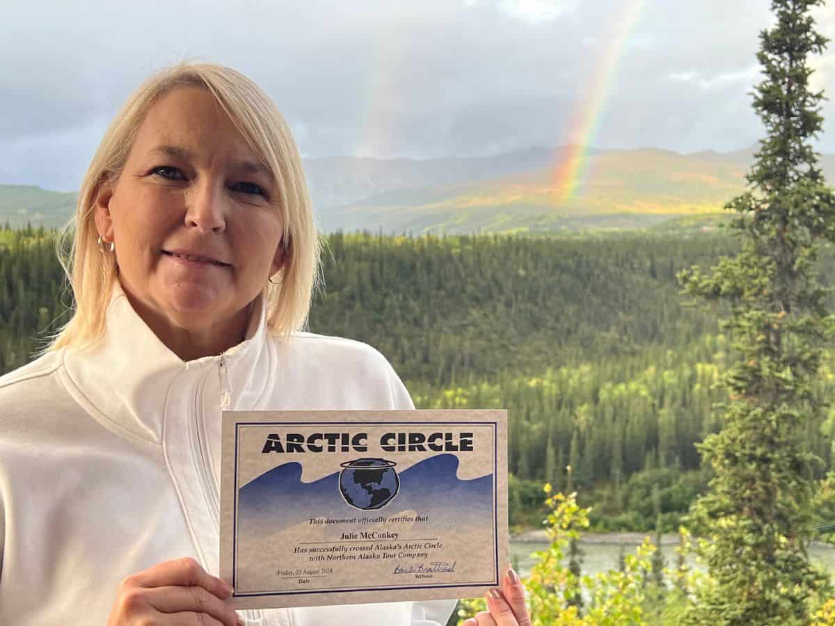 Julie McConkey proudly holds her Arctic Circle certificate from our Denali hotel balcony with rainbows in the background.