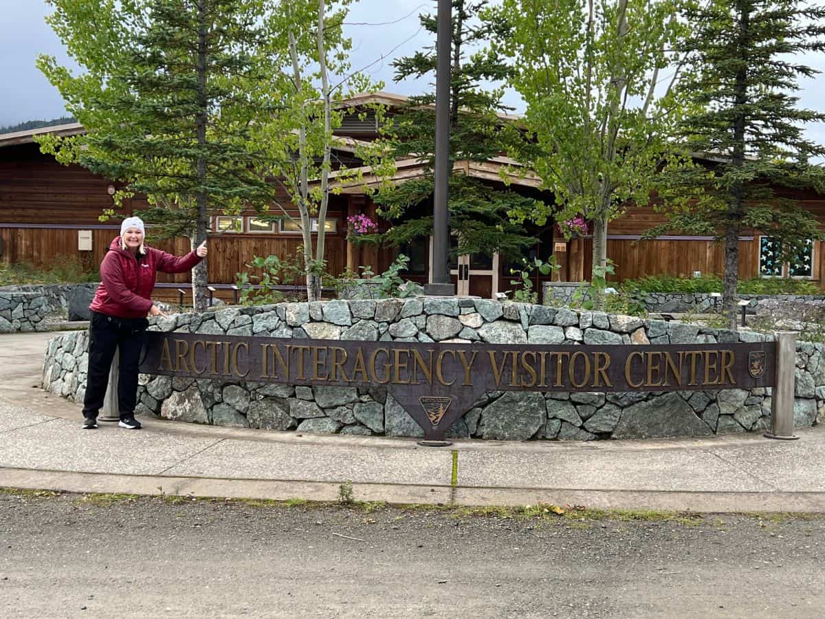 A woman smiles and points to the Arctic Interagency Visitor Center in Coldfoot, AK