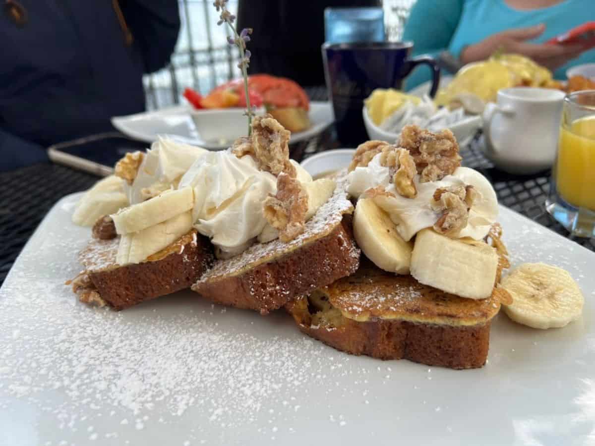 A white plate contains French toast topped with banana slices, nuts, and whipped cream.