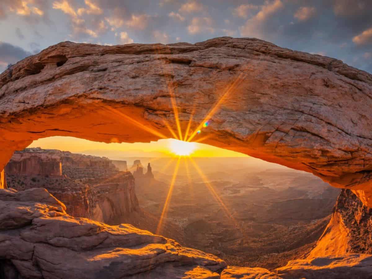 The sun rises above the horizon in the distance as seen below an arch in the foreground.