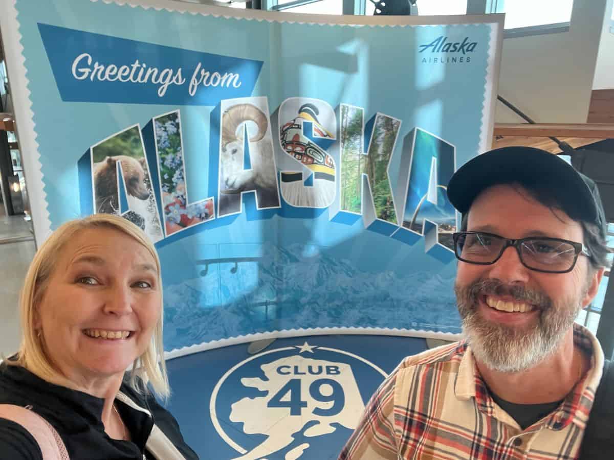 A man and woman pose for a picture in front of the "Greetings from Alaska" sign in the Fairbanks Airport.