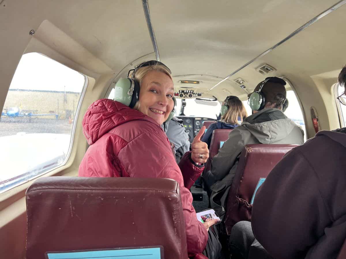 A woman sits in a small 9 passenger plane smiling and giving a thumbs up sign.