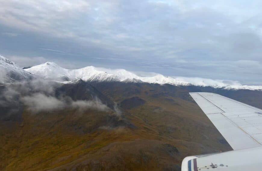 Snowy mountains line the horizon as a plane flies by in the foreground.