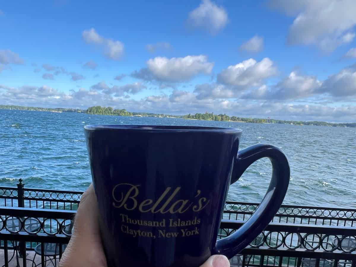 The St. Lawrence River flows in the background behind a hand holding a blue coffee cup.