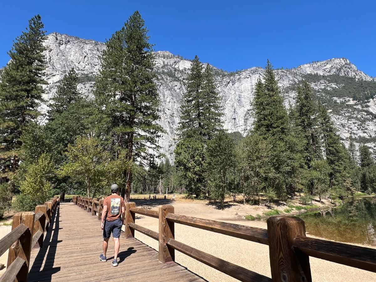 A man walks across a wooden footbridge toward tall trees and granite cliffs.