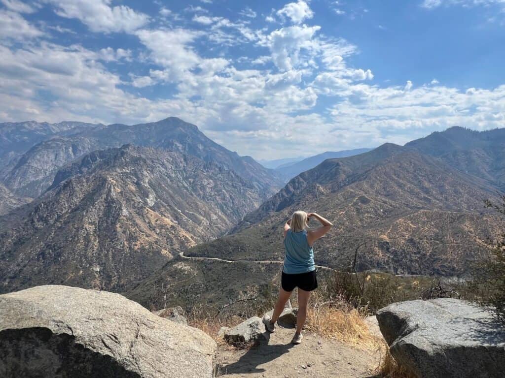 A woman stands at an overlook with a view of a winding road descending between mountains