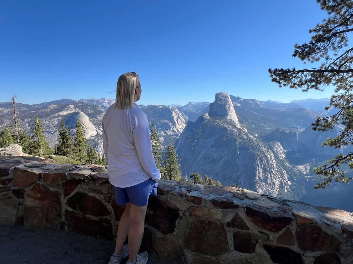 A woman stands behind a stone wall and looks at large granite domes in the distance.