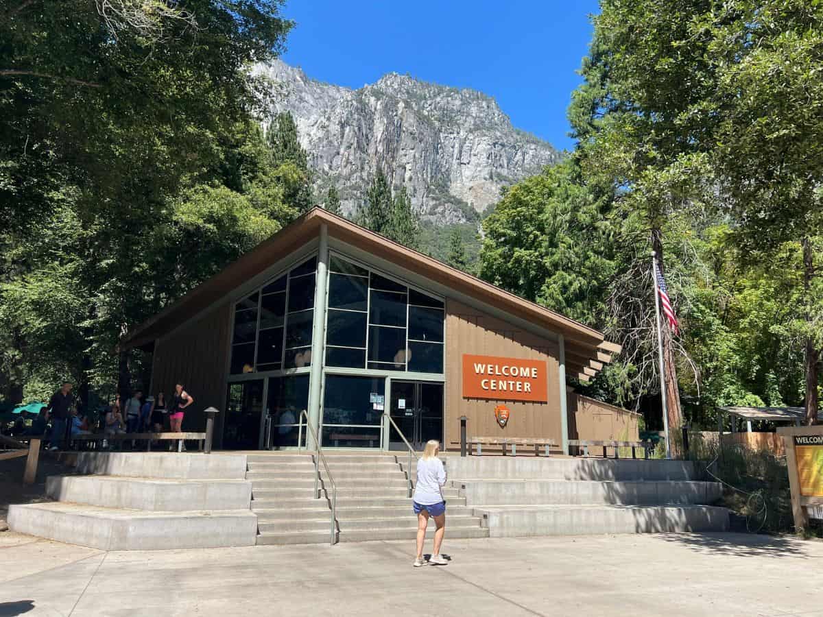 A woman walks toward the Yosemite Valley Welcome Center with a granite mountain behind the building.