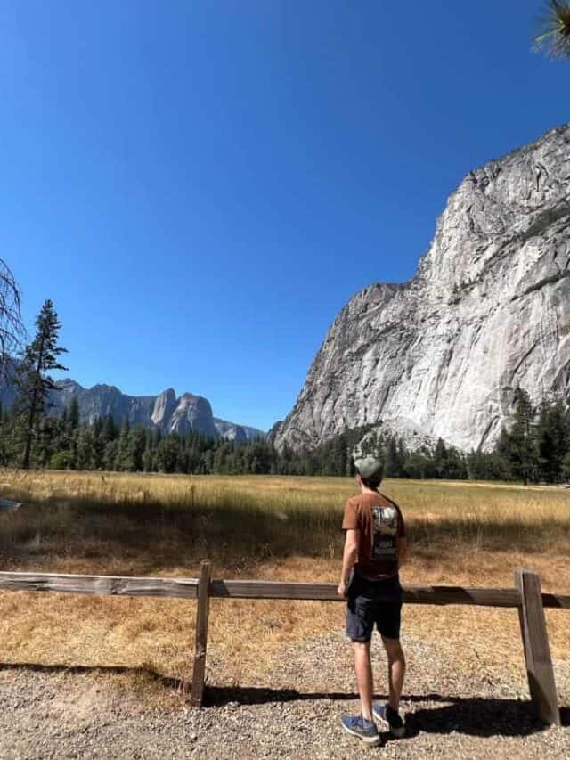 A man stands behind a fence, looking at large granite cliffs across a meadow in Yosemite National Park.