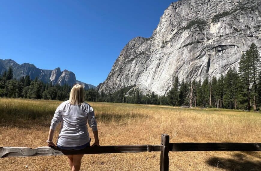 A woman sits on a fence, looking across a meadow at towering granite cliffs.