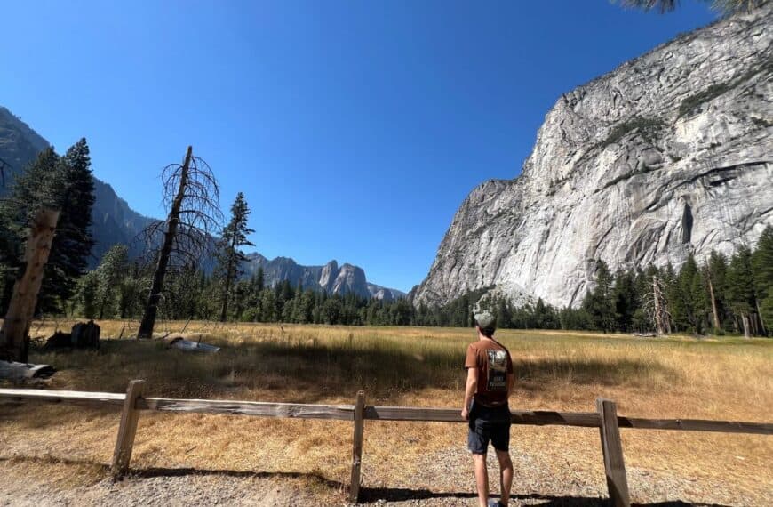 A man stands behind a fence, looking at large granite cliffs across a meadow in Yosemite National Park.
