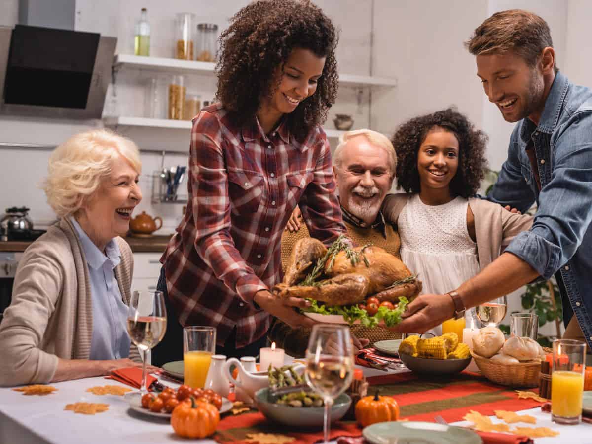 Family members around a table smile as a platter of turkey is set on the table for Thanksgiving dinner.